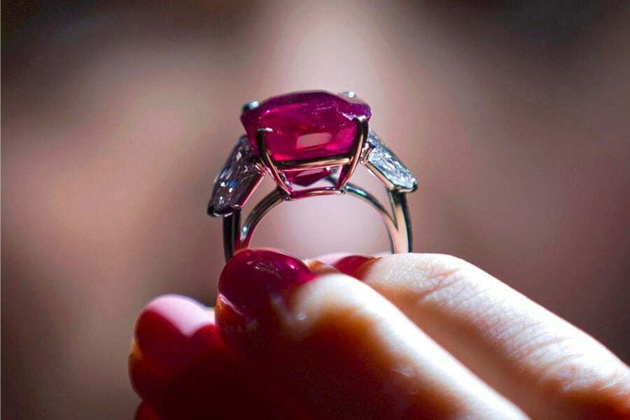 A close up look at the sparkling, pigeon blood red Sunrise Ruby ring held by a hand with pink-colored nail polish