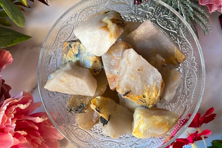 A bunch of raw Rutile crystals in a glass bowl surrounded by flowers and leaves