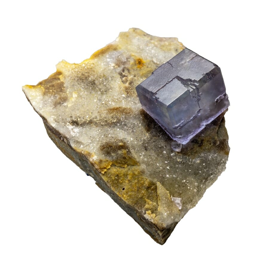 A tiny cube-shaped purple fluorite sitting on top of another pretty mineral