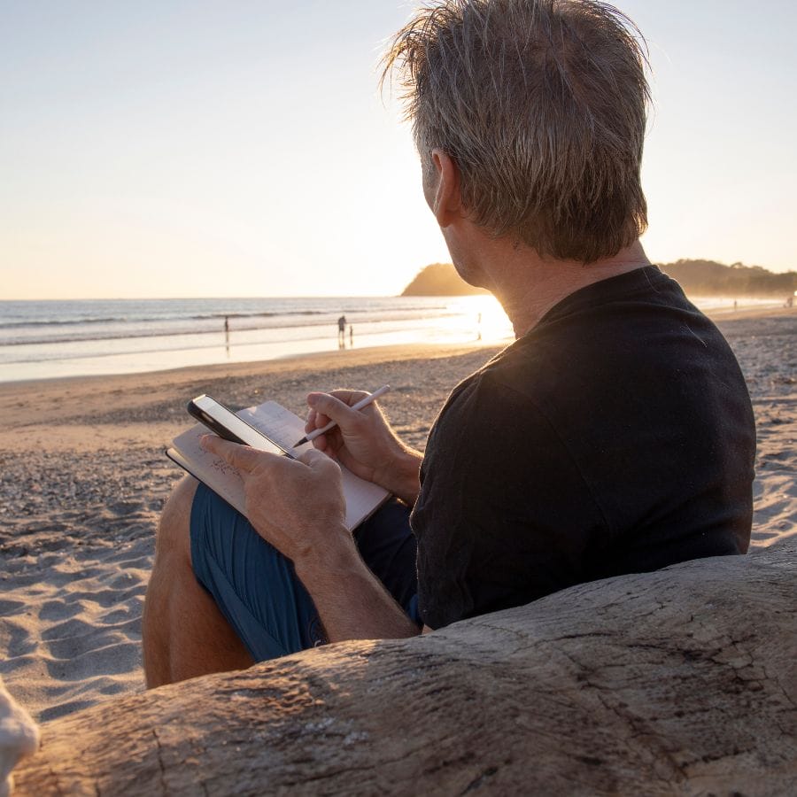 man sitting in a beach and writing in a notebook
